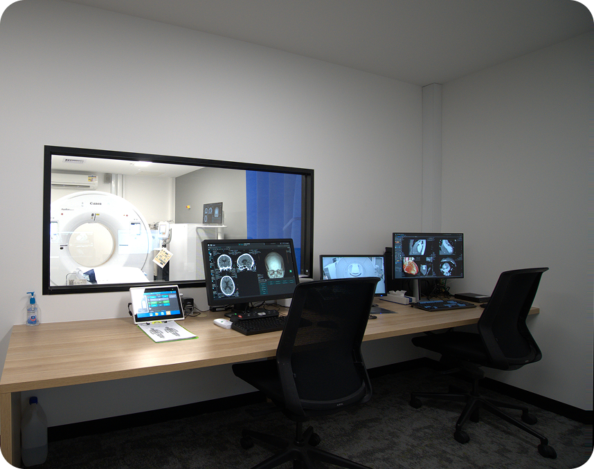 Windowed control room with a CT scanner visible through a large glass window and multiple monitors on a wooden desk in front of black chairs, medical imaging setup.